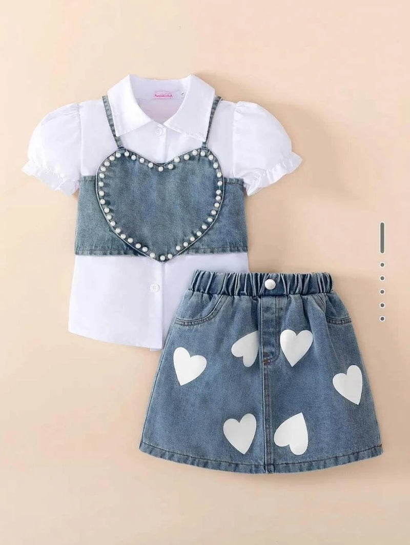 A children's two-piece outfit featuring a white short-sleeve collared shirt with ruffled sleeves and a blue denim mini skirt with white heart patches, laid flat on a light beige background.