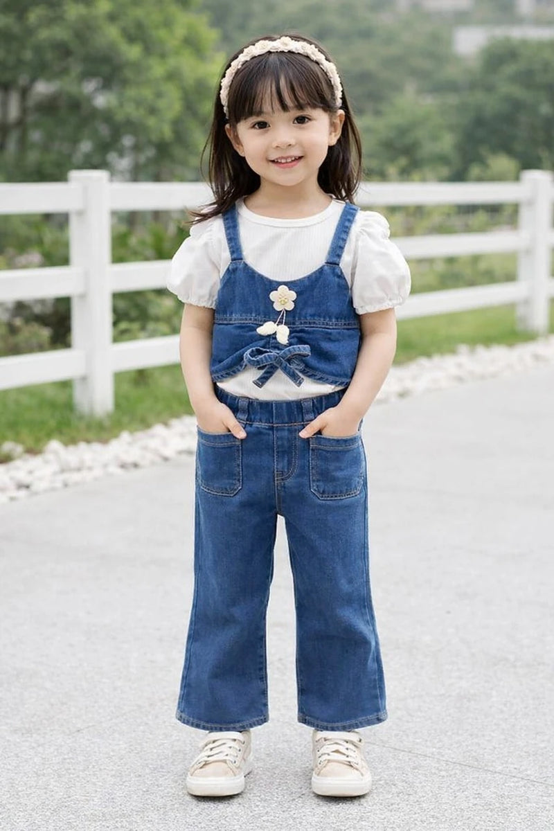 A young girl stands outdoors wearing a blue denim overall-style romper over a white short-sleeve top, with a white headband and white sneakers. The background shows a paved path, a white fence, and green trees.
