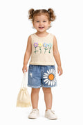 A toddler girl with curly hair in pigtails stands against a white background, wearing a cream sleeveless top with embroidered flowers and blue denim shorts with a daisy patch. She is holding a small cream fringed drawstring bag.