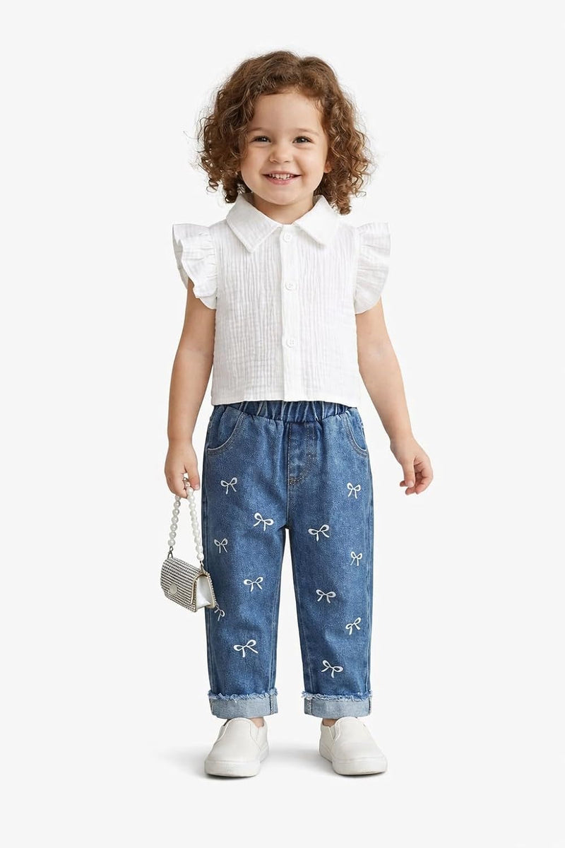 A toddler girl with curly brown hair stands against a light gray studio background wearing a white sleeveless collared top with ruffled shoulders, blue denim pants with small white bow prints, and white shoes. She is holding a small metallic handbag.